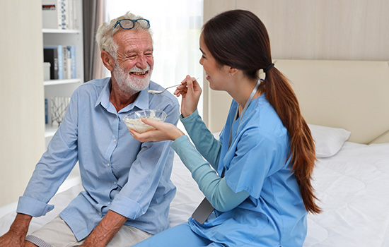Happy caregiver sitting and feeding senior caucasian man on bed with rice porridge soup for breakfast at retirement house. Asian smiling nurse taking good help care and support elder patient at home.